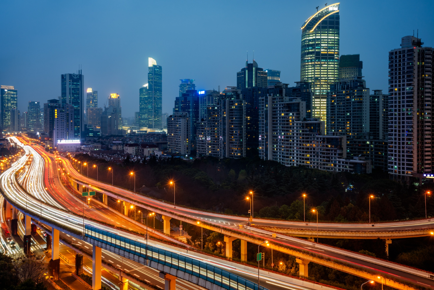 Night cityscape with illuminated skyscrapers and highways showing light trails from moving vehicles.