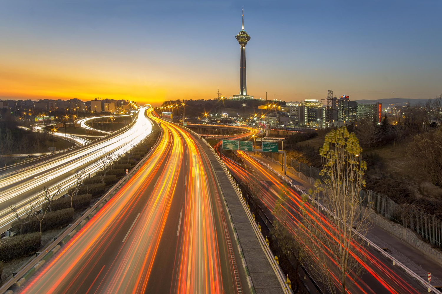 Long-exposure view of busy highway with light trails near a tall tower during sunset in a city.