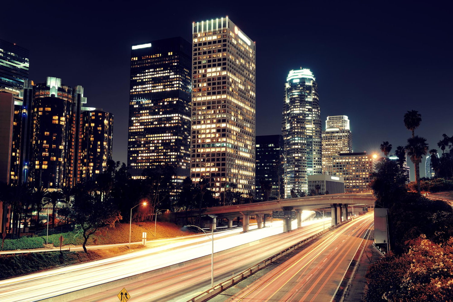 Night cityscape with tall illuminated skyscrapers and light trails on busy highway below.