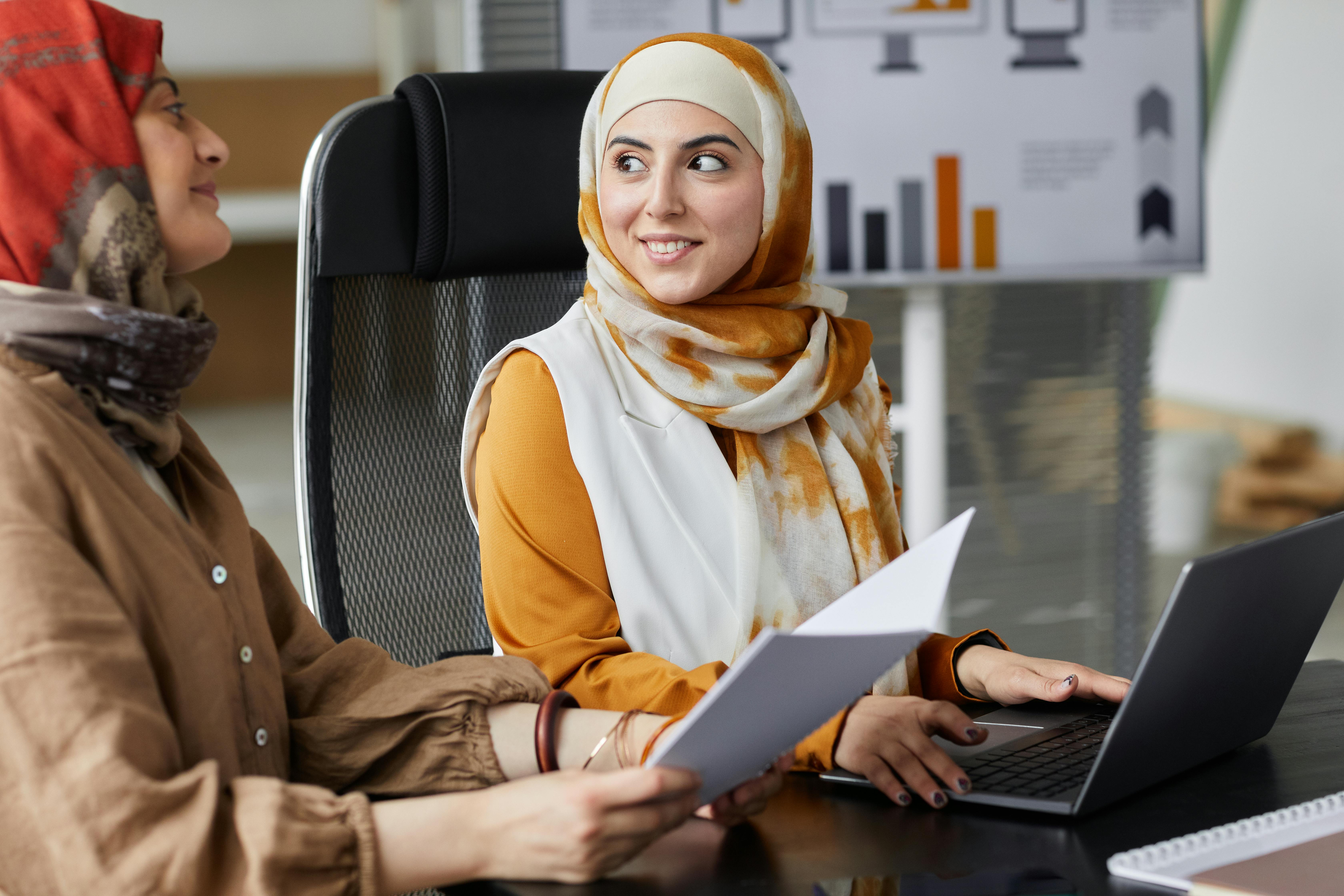 Two employees looking at each other while sitting at a desk with a laptop and paper