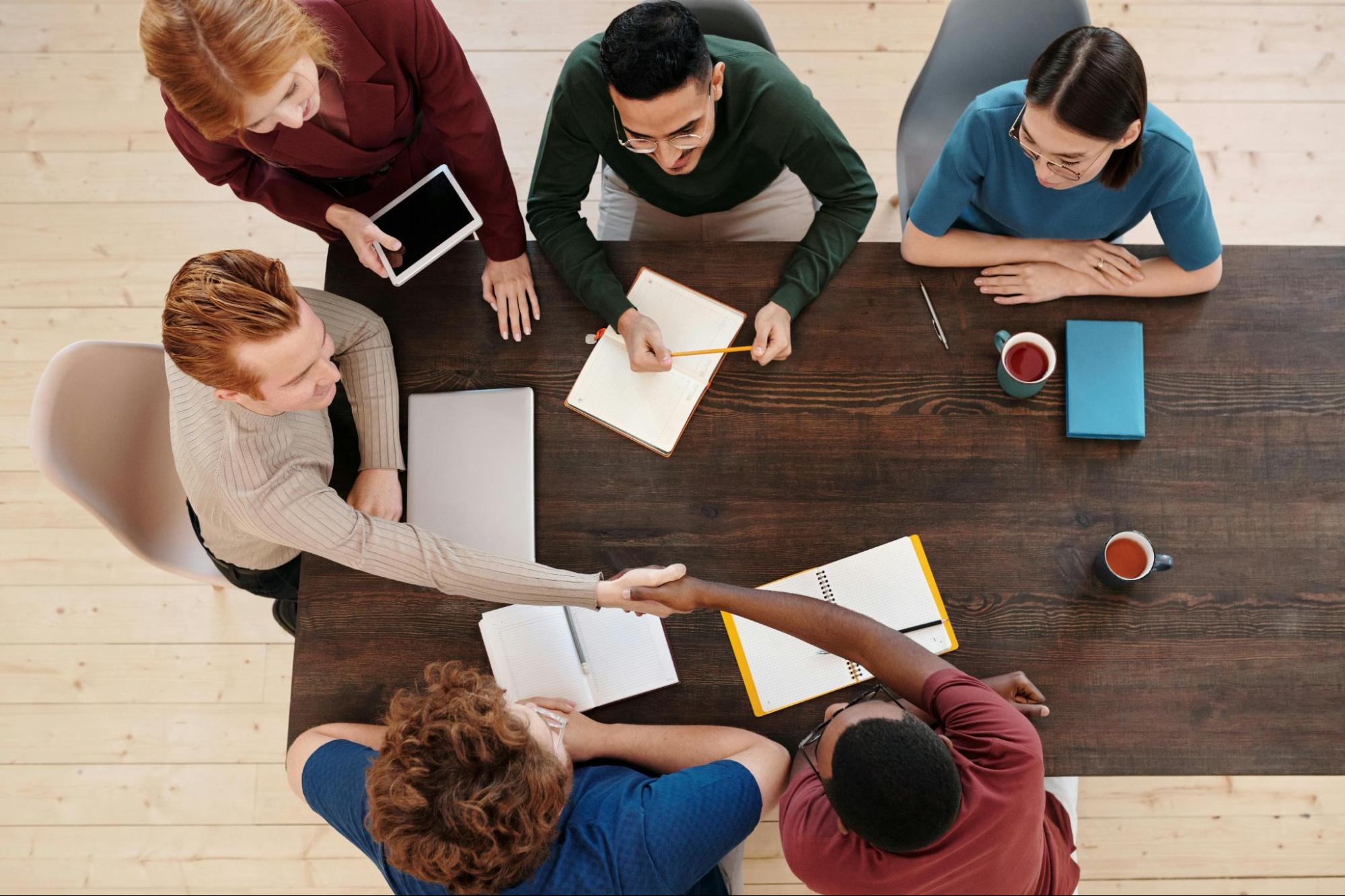 Six co-workers sit around a table with notepads and coffee, with two people shaking hands.