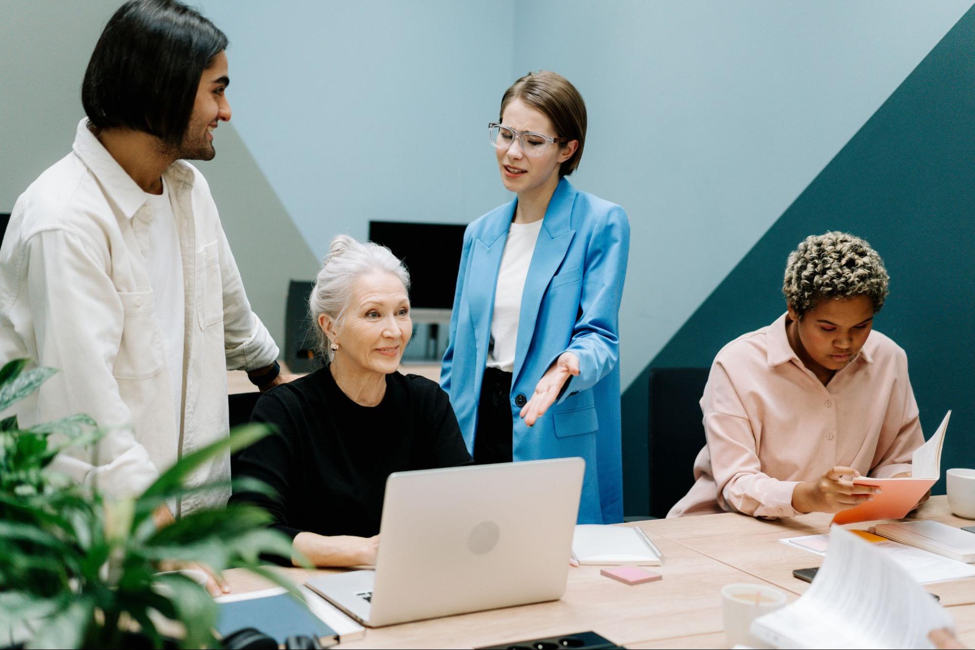 A group of four people, one using a laptop at a desk, two standing over the laptop gesturing to it, and another sitting and reading a booklet off to the side.