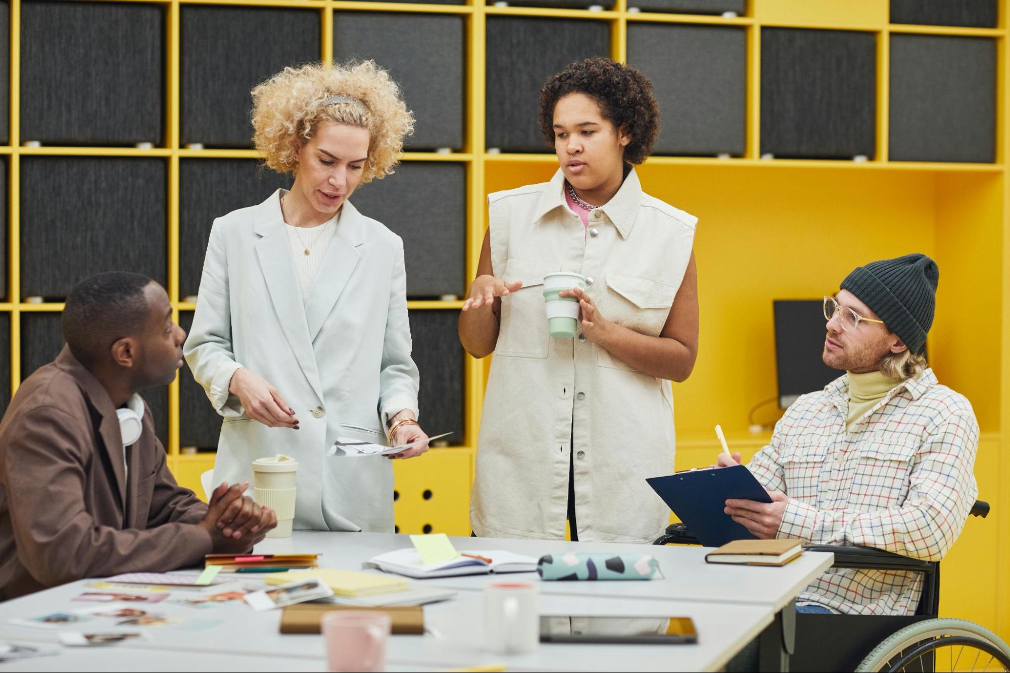Four people (two sitting, two standing) gather around a table talking and taking notes.