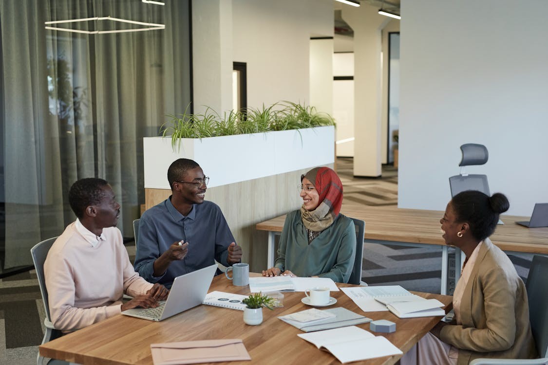 Four people holding a discussion over a desk, with notebooks and a laptop.