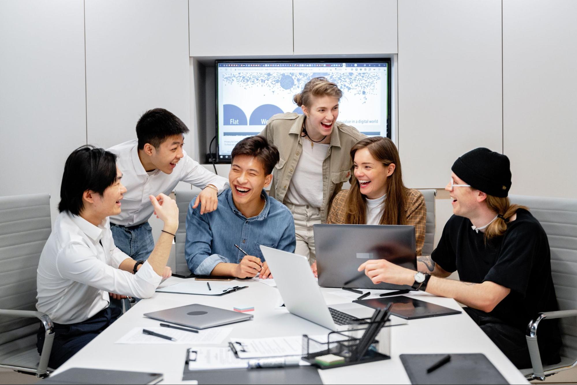 A team of six people laughing and celebrating around a table with laptops on it.