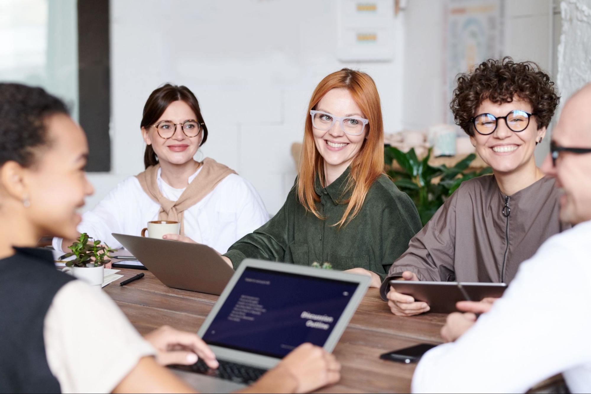 Five seated people smile at each other at a table with laptops and tablets on it. 