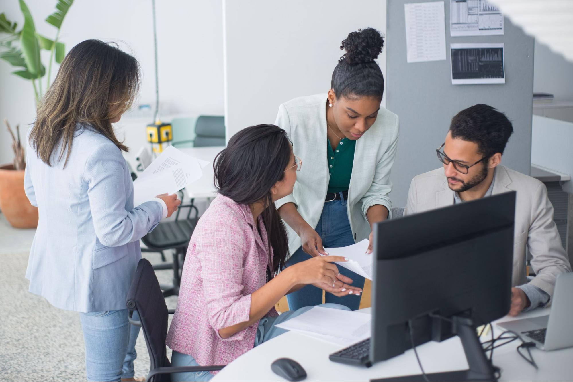 Four employees in an office setting, discussing paperwork by a computer.