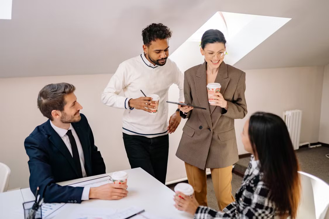 Four people having a discussion at a desk over coffee and a tablet.