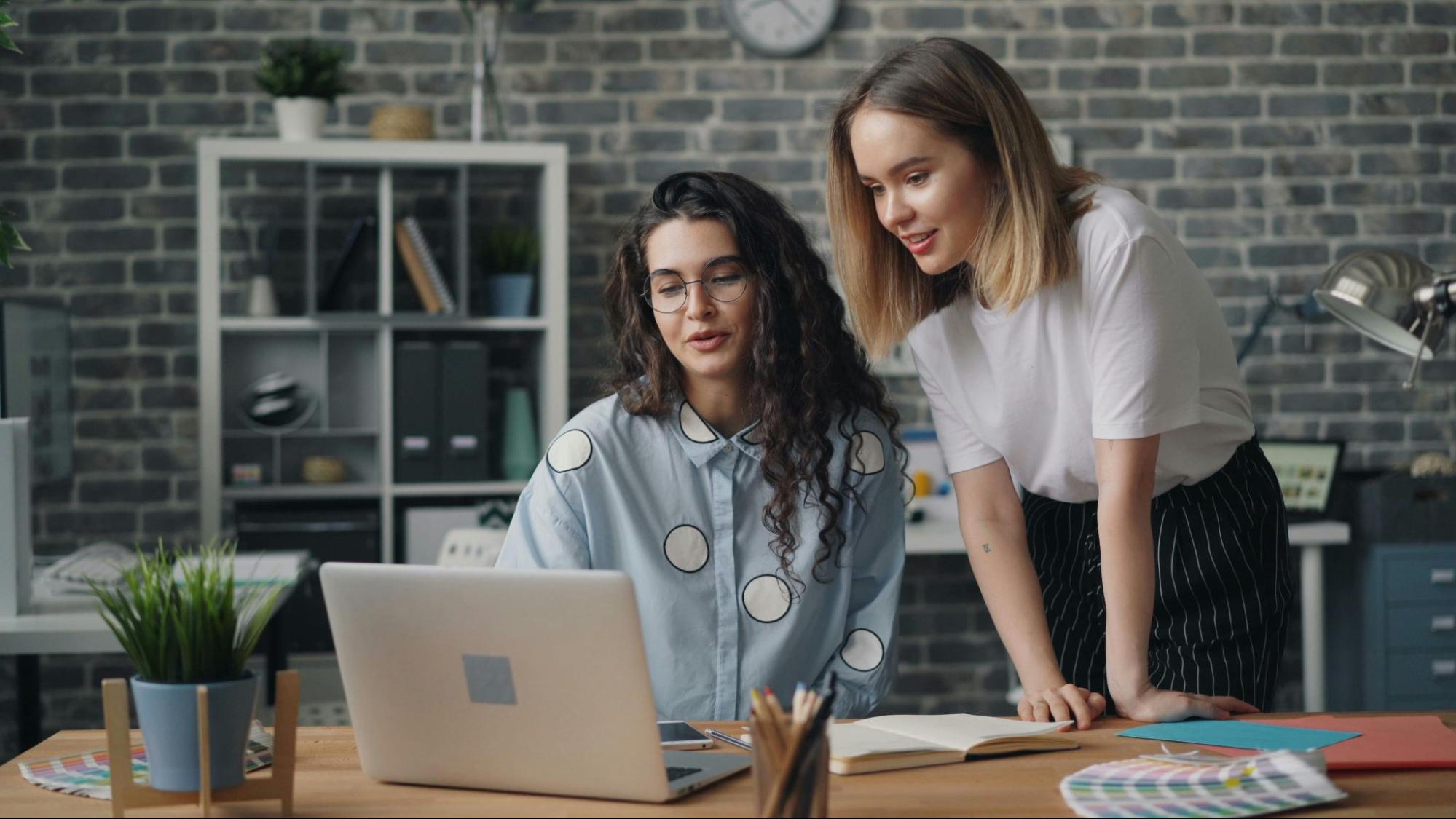 Two people look at a laptop on a desk with paperwork, a jar of pens, and a plant.