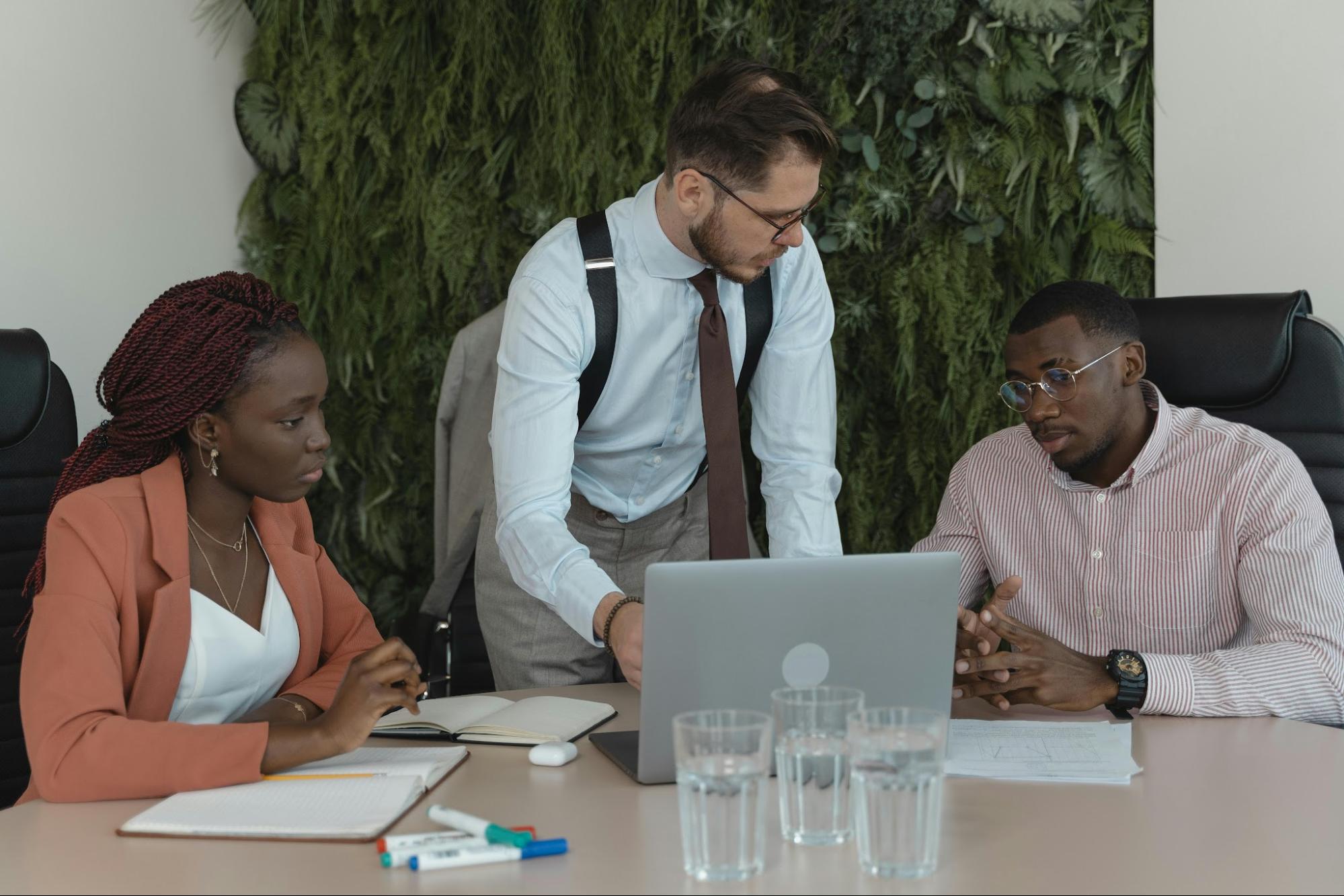 Three employees discuss and look at a laptop on a table alongside notebooks, markers, and glasses of water.