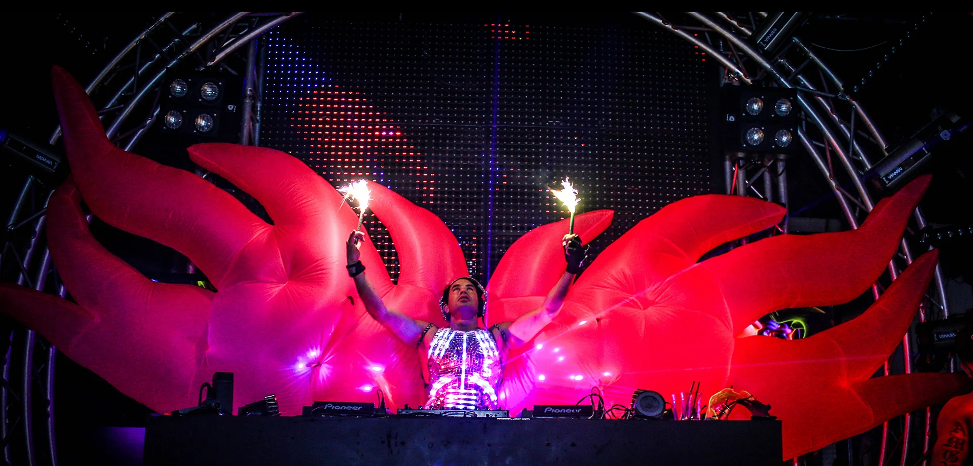 Kidd Madonny with inflatable red flame wings at a club, holding sparklers above his head behind a DJ booth.