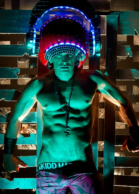 Kidd Madonny wearing large glowing helmet and patterned shorts in neon-lit setting.