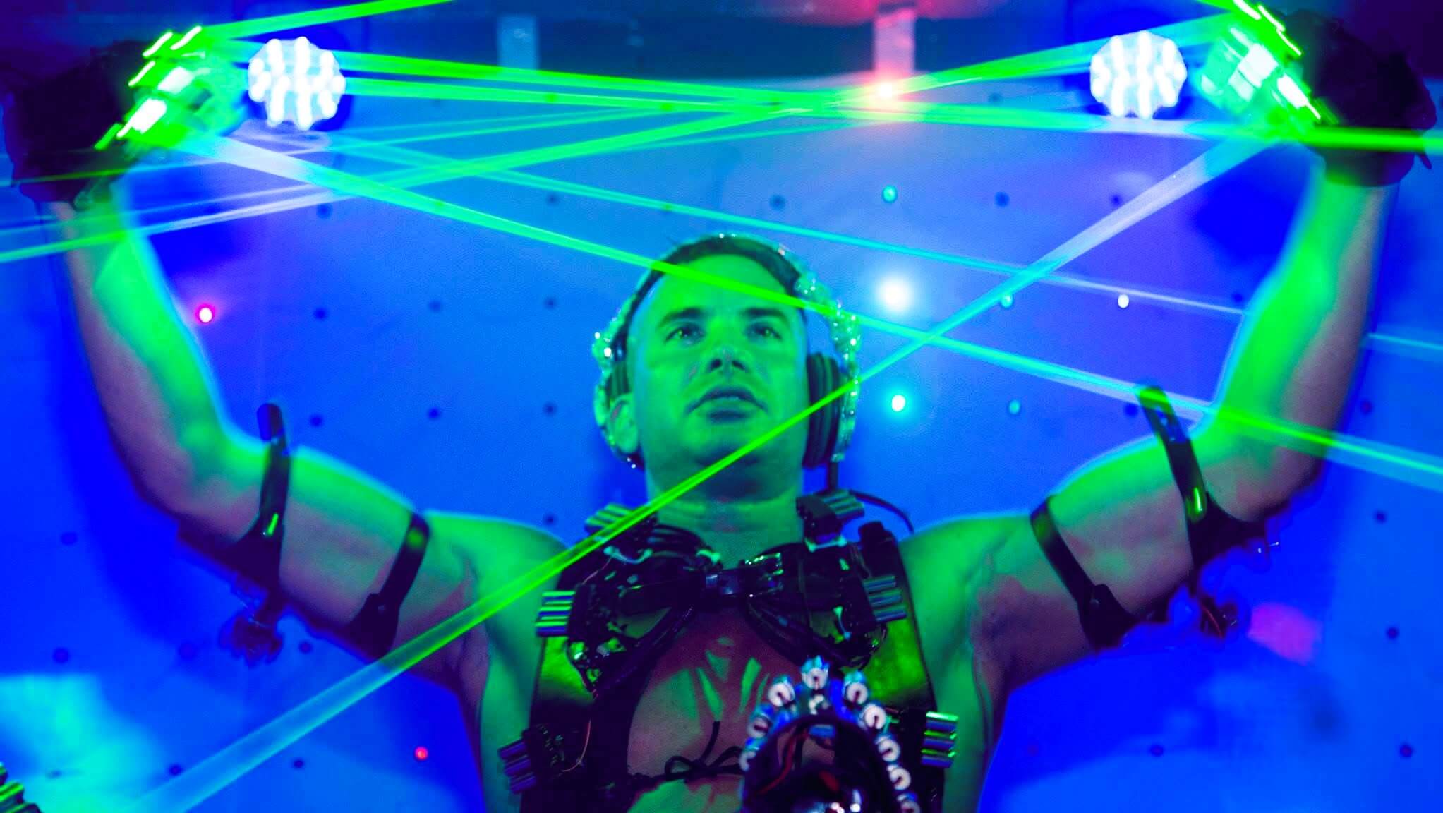 Kidd in black harness and headphones manipulating green laser beams against a blue background.