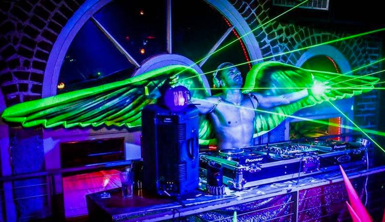 Kidd Madonny DJ with green illuminated wings stands behind a mixing console shooting green laser beams.