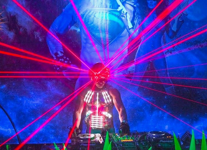 Kidd Madonny performing with red laser lights and neon body paint against a blue superhero mural background.