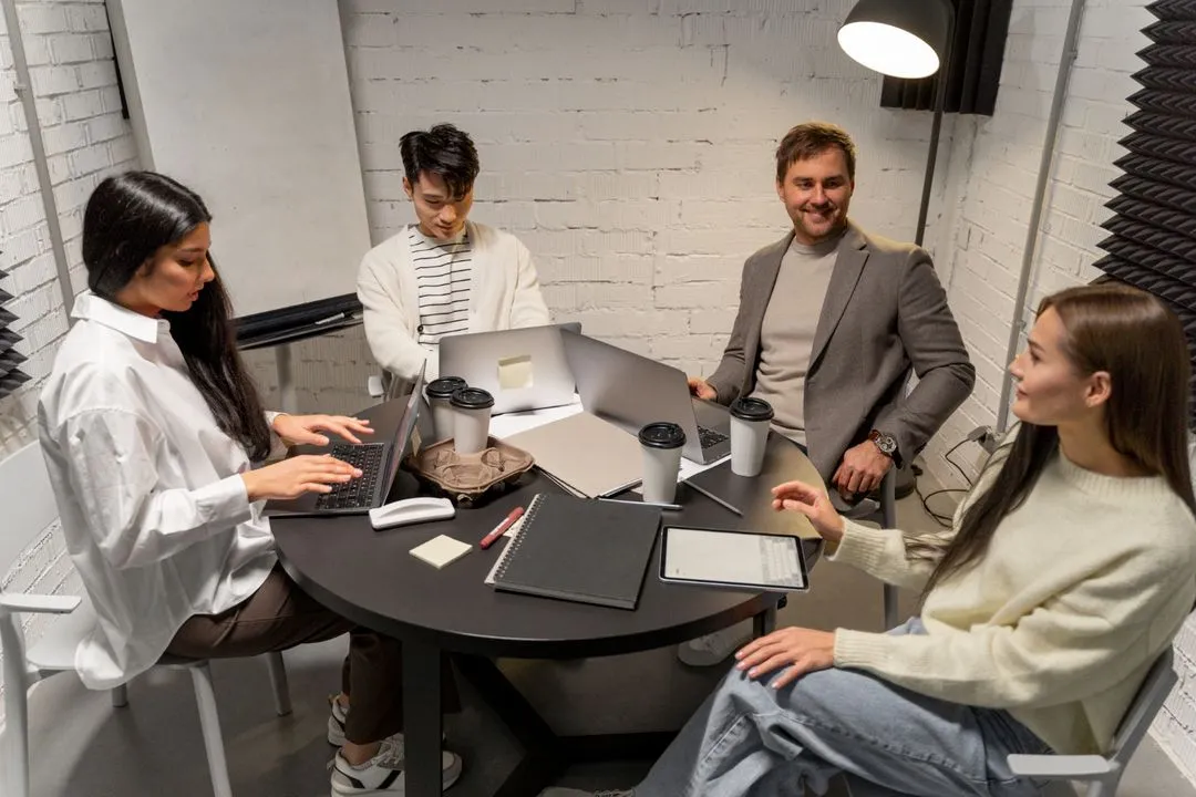 Four diverse colleagues at a round table in a modern office, working on laptops and discussing.