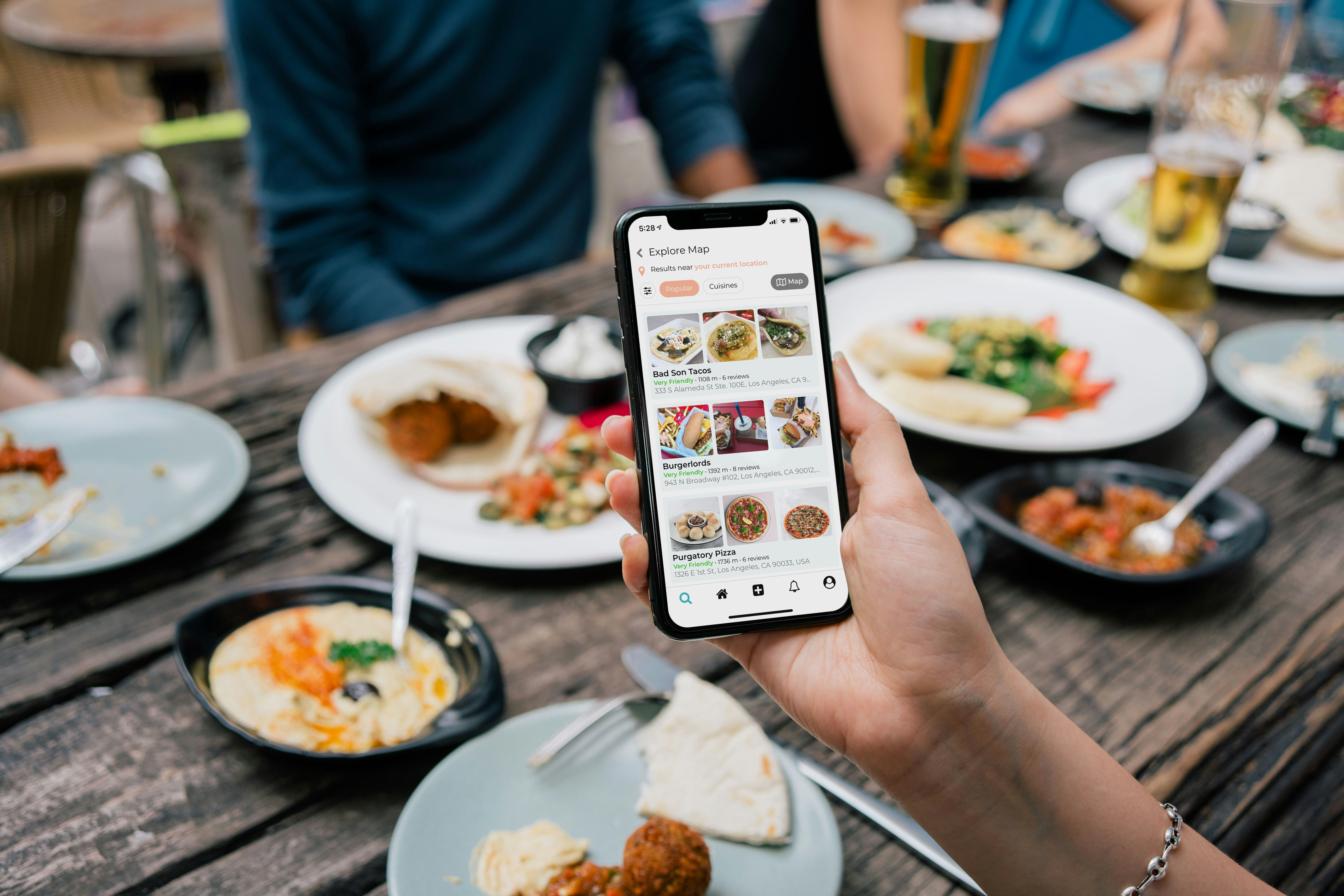 Hand holding phone showing food app with restaurant options over table with various dishes and drinks.