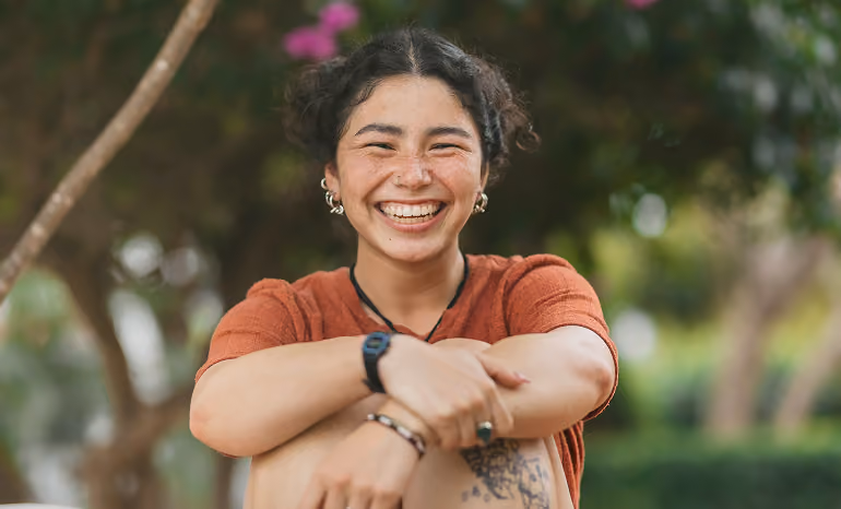 Smiling young woman with curly hair, freckles, and earrings sitting outdoors with arms wrapped around knees.