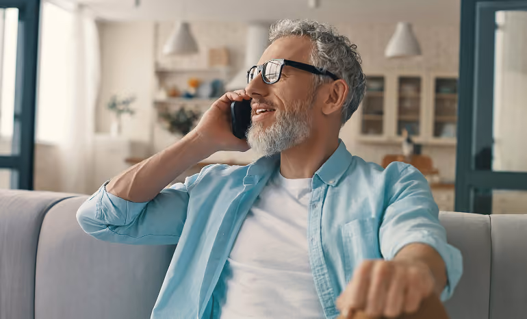 Smiling man with gray hair and beard talking on phone while sitting on a couch in a bright room.