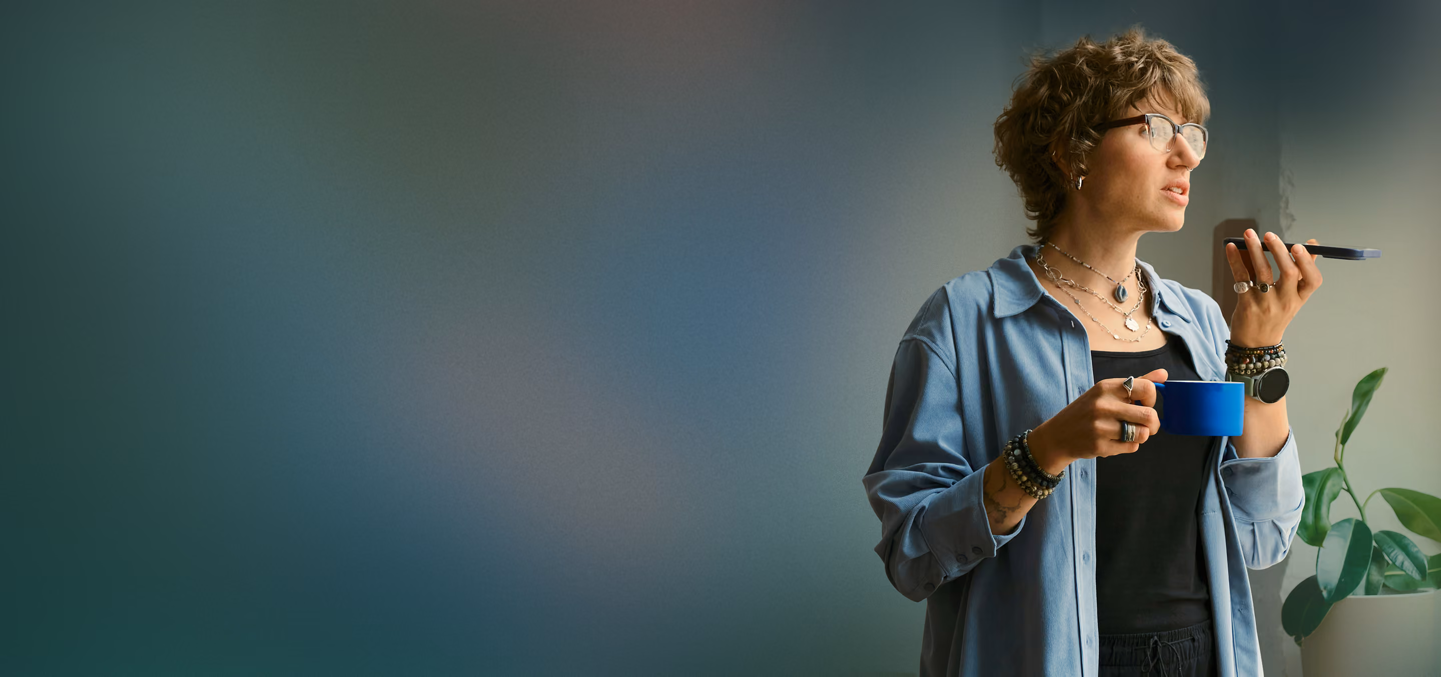 Young person holding a blue mug and speaking into a smartphone, standing near a potted green plant.