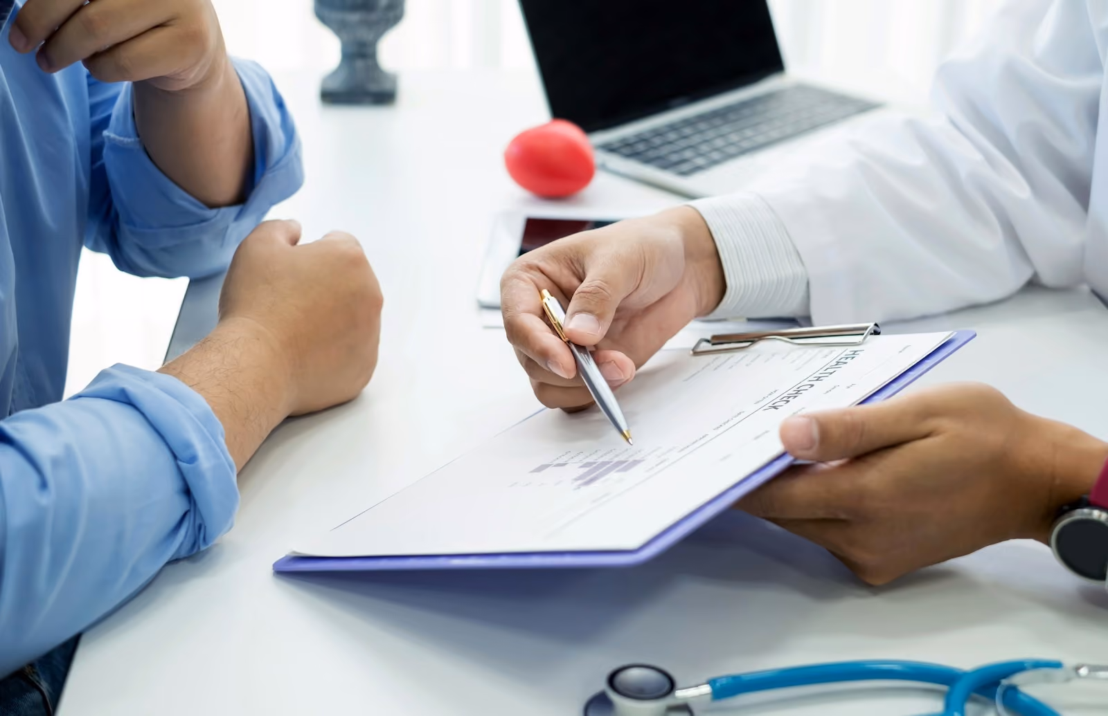Doctor in a white coat reviewing a clipboard chart with a seated patient in a blue shirt