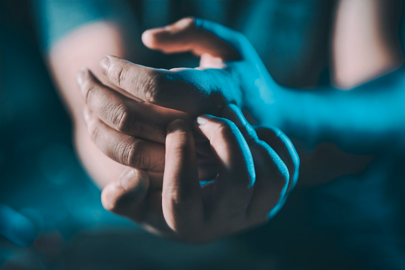 Close-up of two clasped hands lit in teal tones, one gently rubbing the other