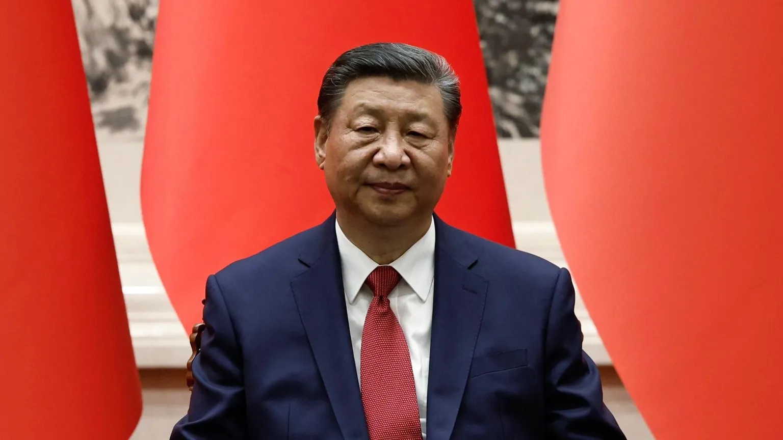 Chinese political leader seated in front of large red flags.