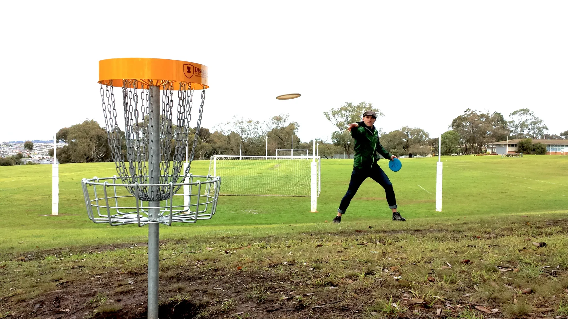 A player putting toward an orange-topped disc golf basket on an open grassed oval, a disc in mid-flight