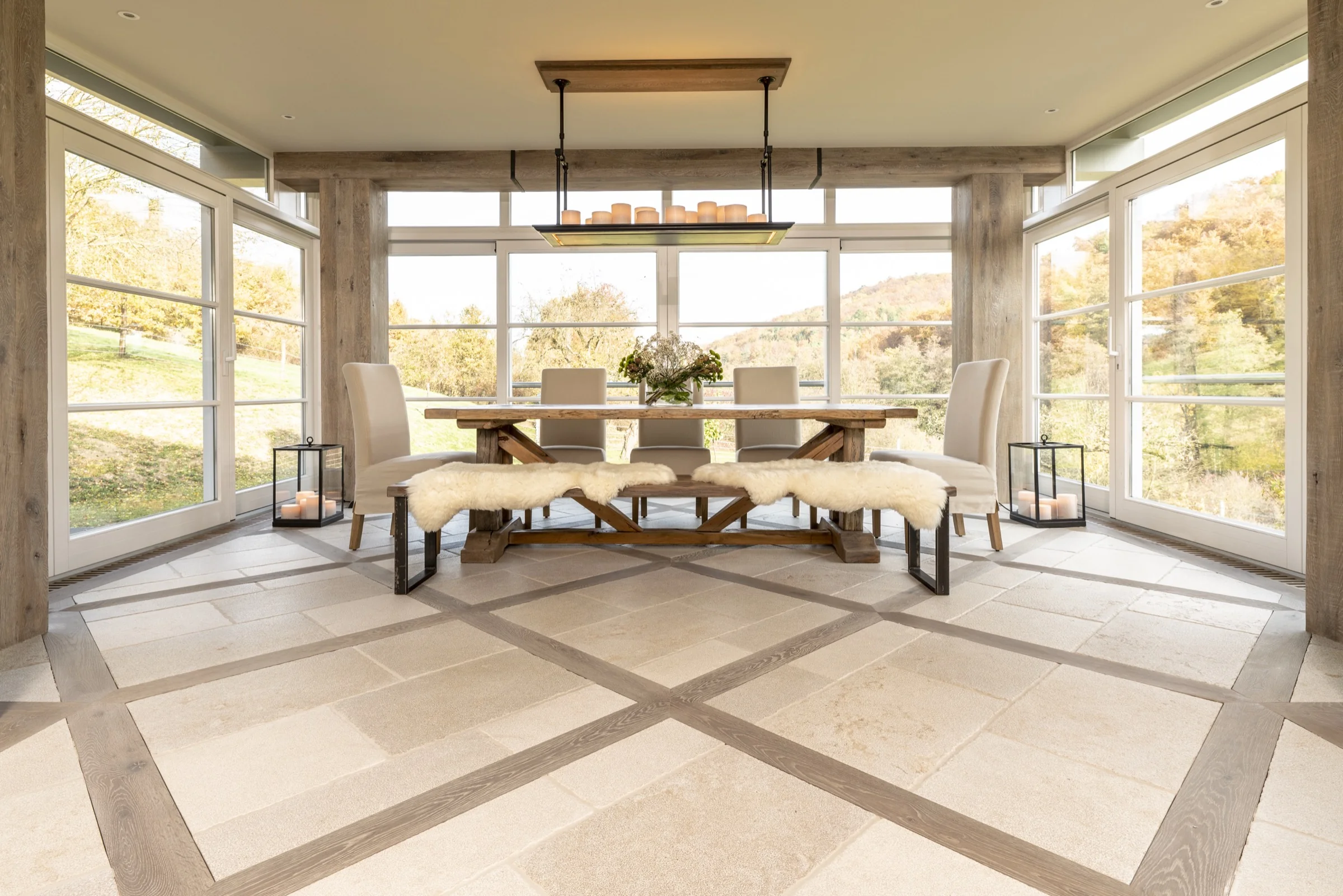 Bright dining room with large windows showing an outdoor autumn landscape, featuring a wooden table, three beige chairs, two benches with white fur covers, and candle lanterns on the floor near the windows.