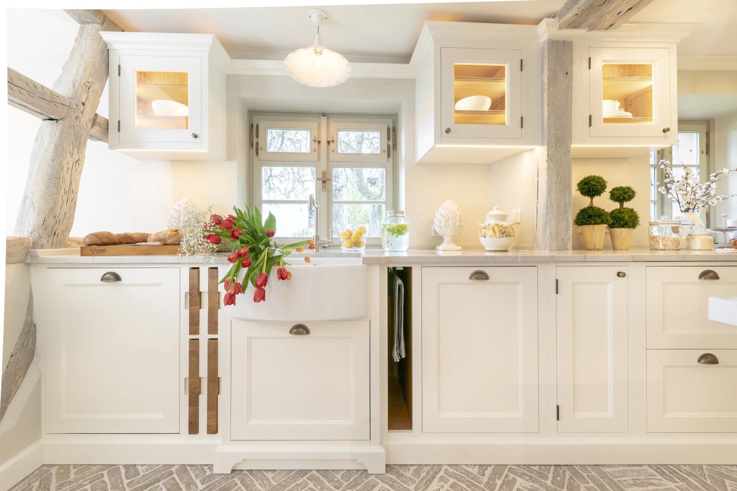 Bright white kitchen with farmhouse sink below a window, red tulips on the sink, wooden bread board with bread, decorative jars, and green topiary plants on the countertop.