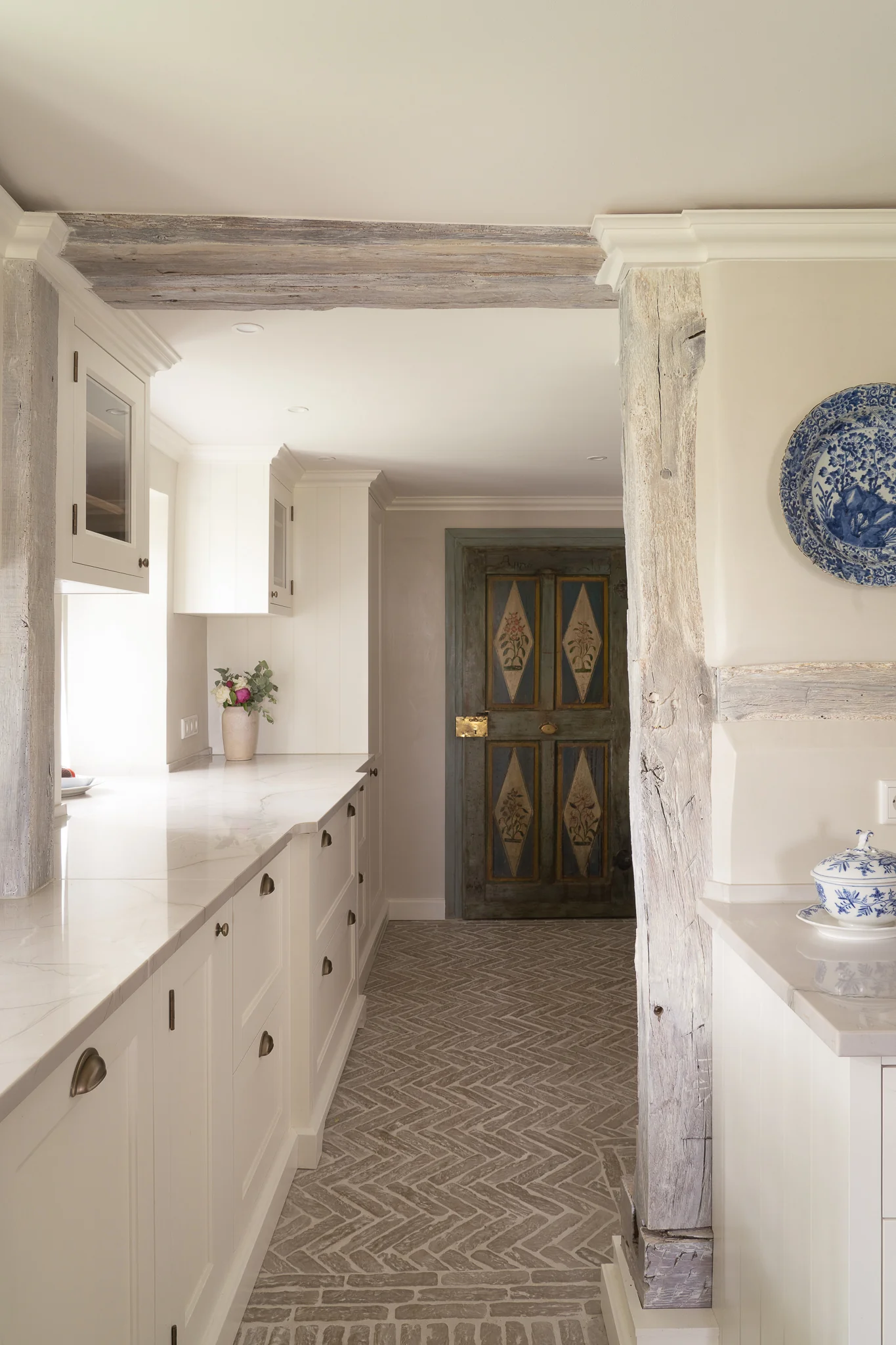 Narrow kitchen corridor with white cabinetry and marble countertops, rustic wooden beams, patterned stone floor, decorated blue and white plate on the wall, and a door at the end with floral diamond panels and brass hardware.