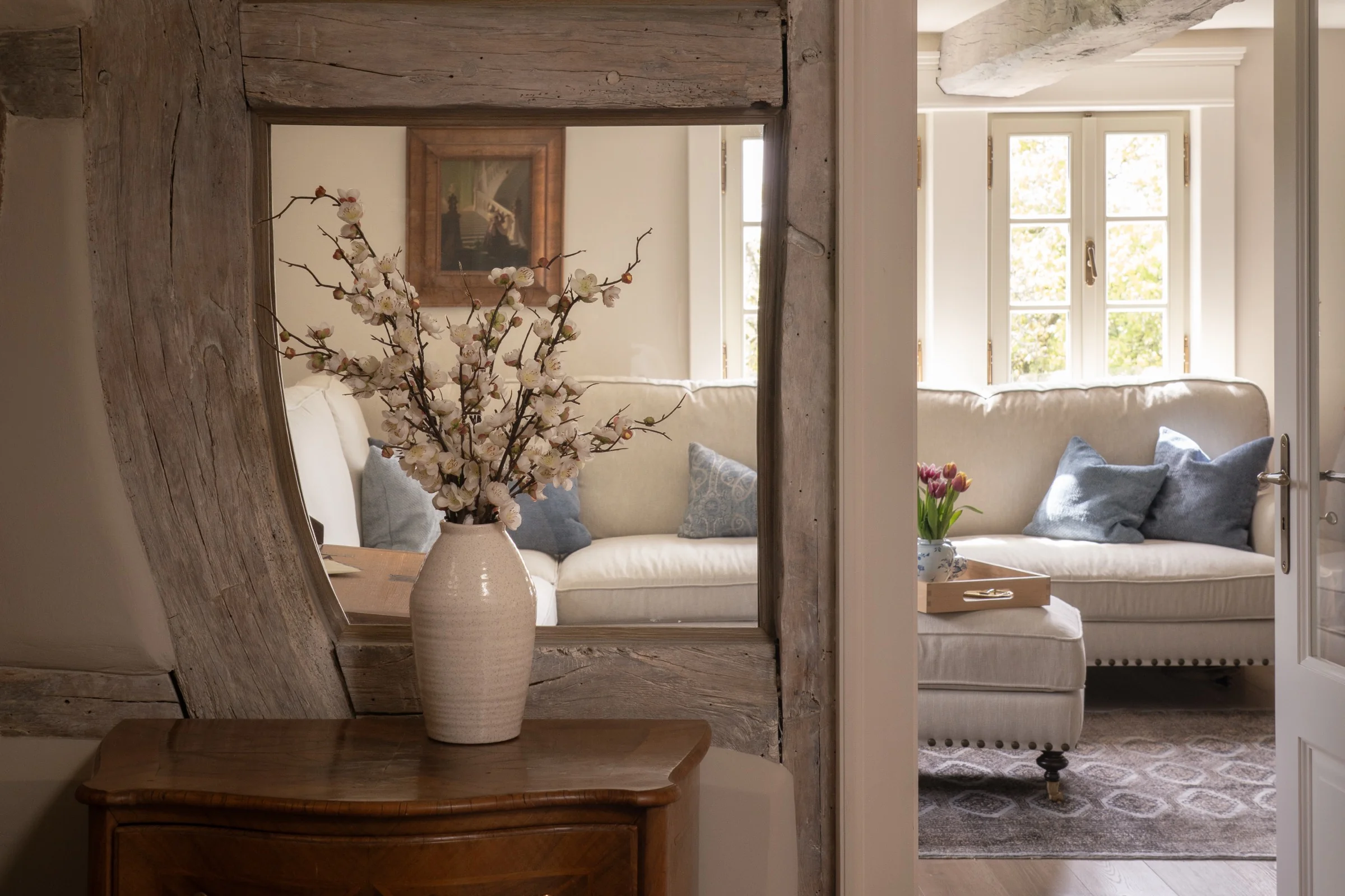 Cozy living room with a large beige sectional sofa adorned with blue throw pillows, a vase of white flowering branches on a wooden side table in front of a rustic wooden-framed mirror, and a window letting in natural light.