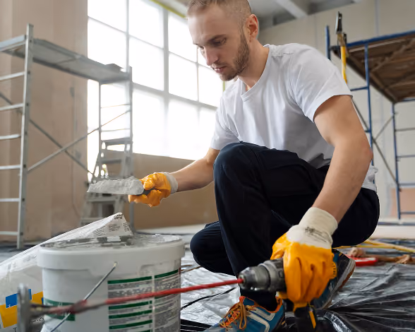 Man in orange gloves mixing paint with drill in a bright room under renovation.