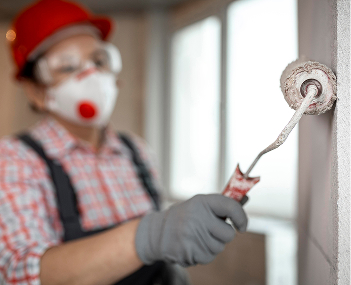 Worker wearing mask and gloves painting a wall with a roller in a bright room.