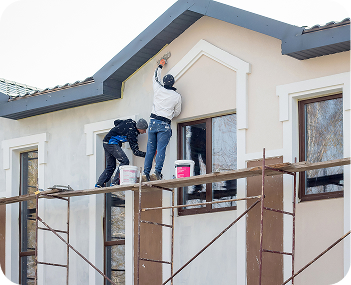 Two workers on scaffolding painting the exterior wall of a beige house under the roof eaves.