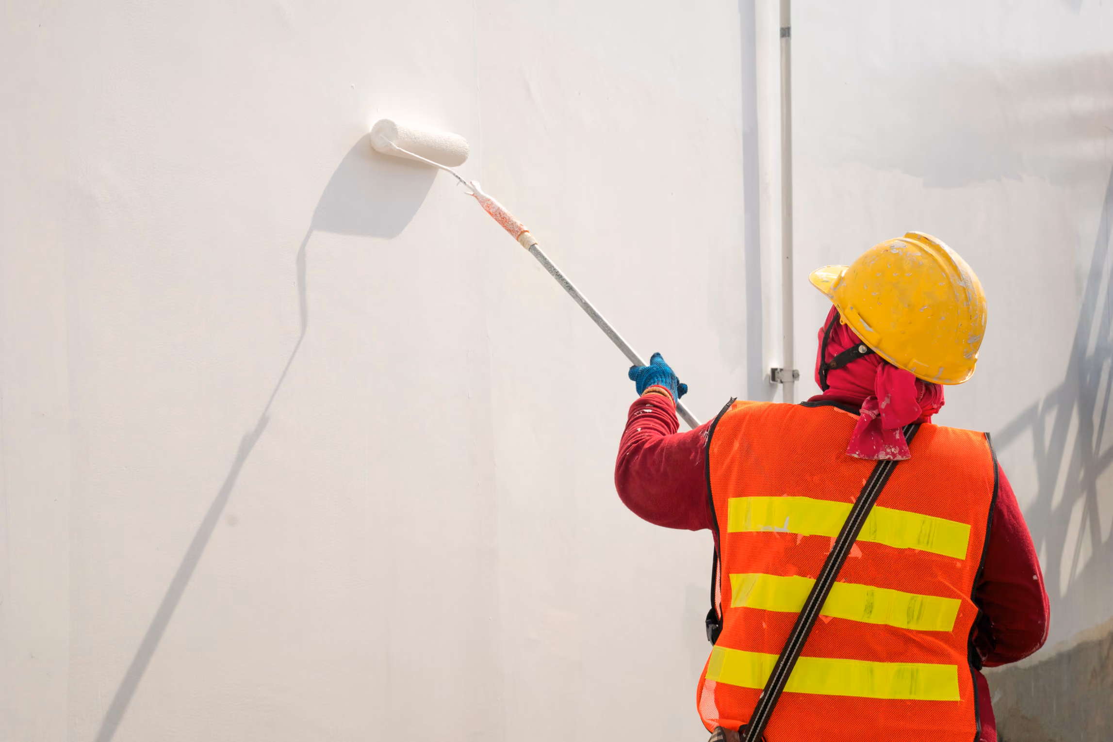 Worker in yellow helmet and orange safety vest painting a white wall with a roller brush.