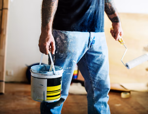 Person wearing paint-stained jeans holding a paint bucket and a roller in a room under renovation.