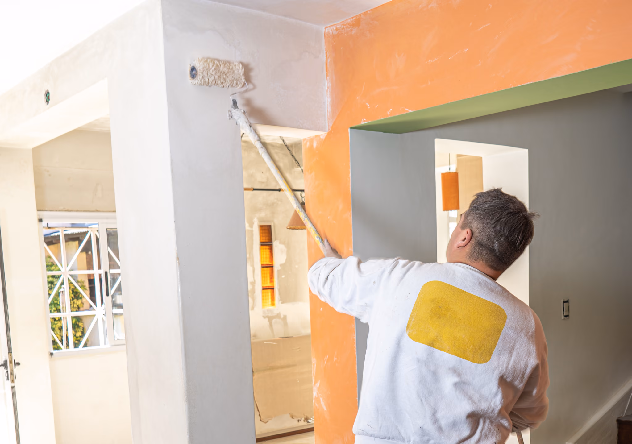 Man painting an interior wall white with a roller on an extension pole near an orange wall.