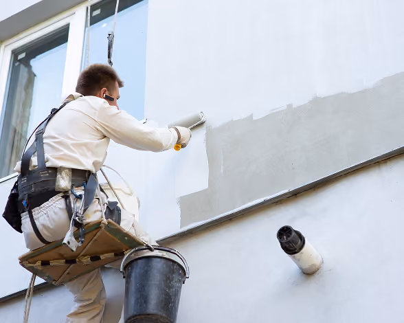 Worker wearing safety gear painting a building wall white while suspended on a platform.