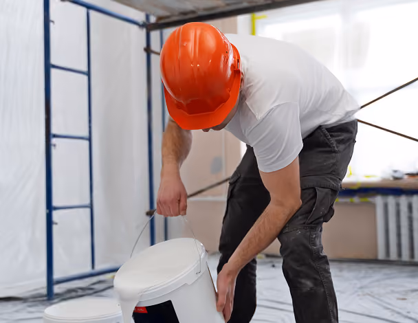 Construction worker wearing an orange hard hat bending down to pick up a white bucket indoors.