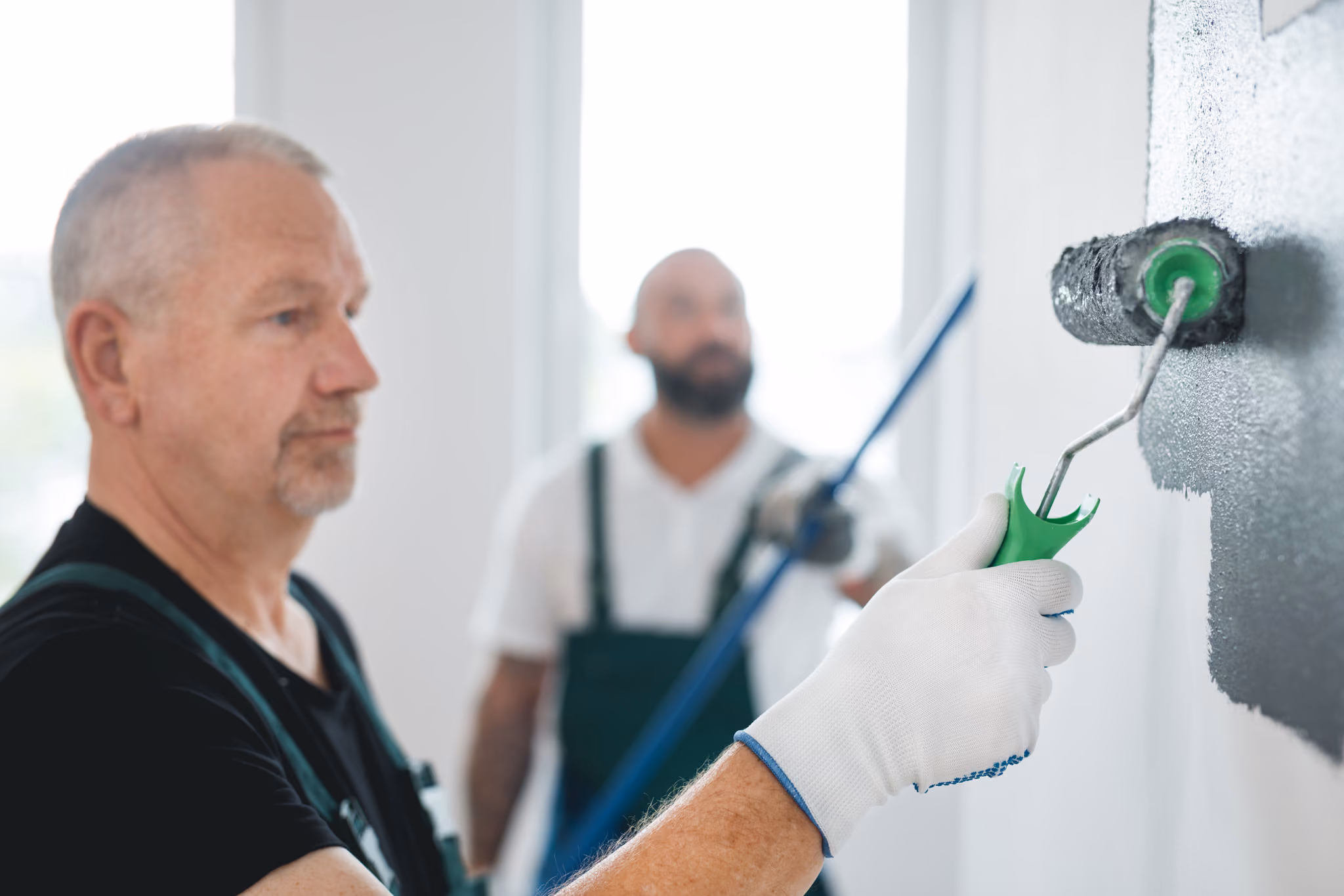 Close-up of a man in gloves painting a white wall black with a paint roller, another man in background.