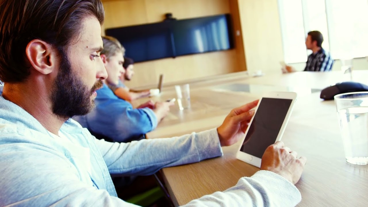 Participants at a conference table with a tablet, wall-mounted displays and a conference phone.