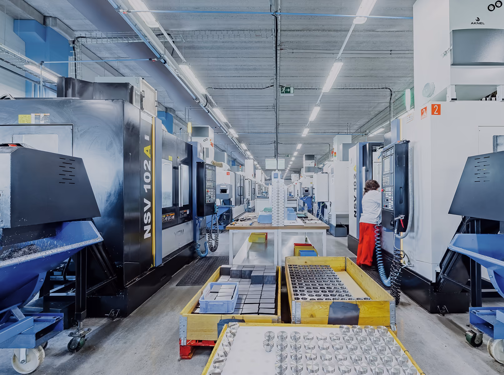 Moderne Fabrikhalle mit CNC-Maschinen, Werkstücken auf Paletten und Arbeiter an Steuerung.