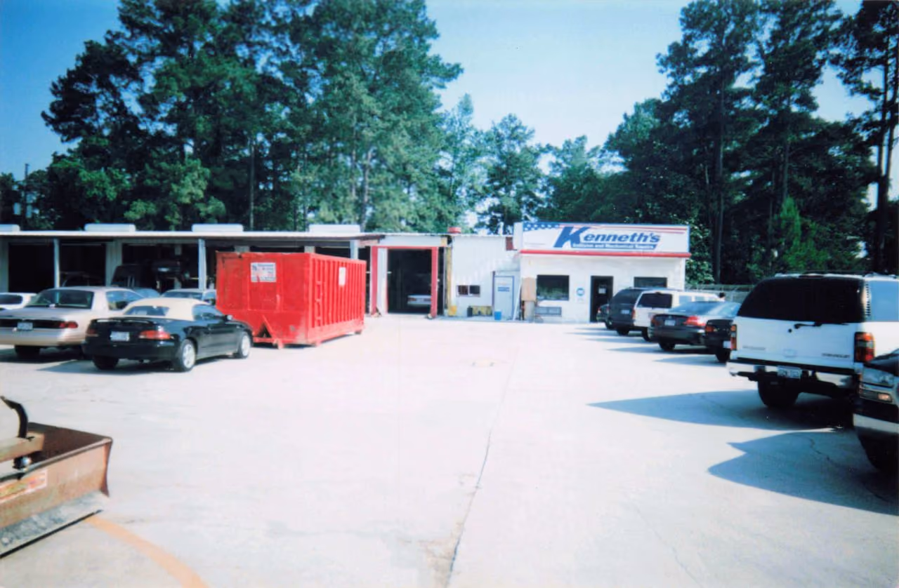 A wide shot of Kenneth's Car Care auto repair shop showing parked vehicles, garage bays, a red dumpster container, and a sign reading "Kenneth's Collision and Mechanical Repairs."