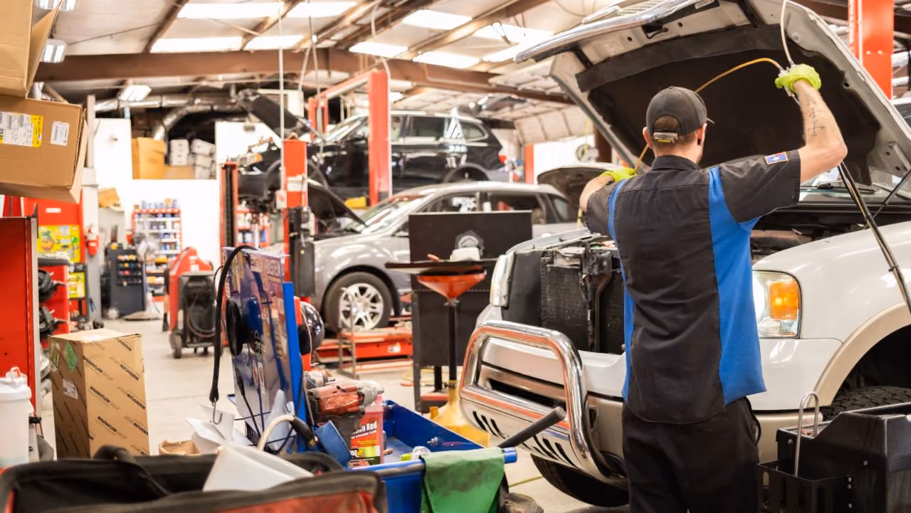 A mechanic viewed from behind works under the hood of a white truck using specialized fluid equipment in a busy garage filled with tools and elevated vehicles.