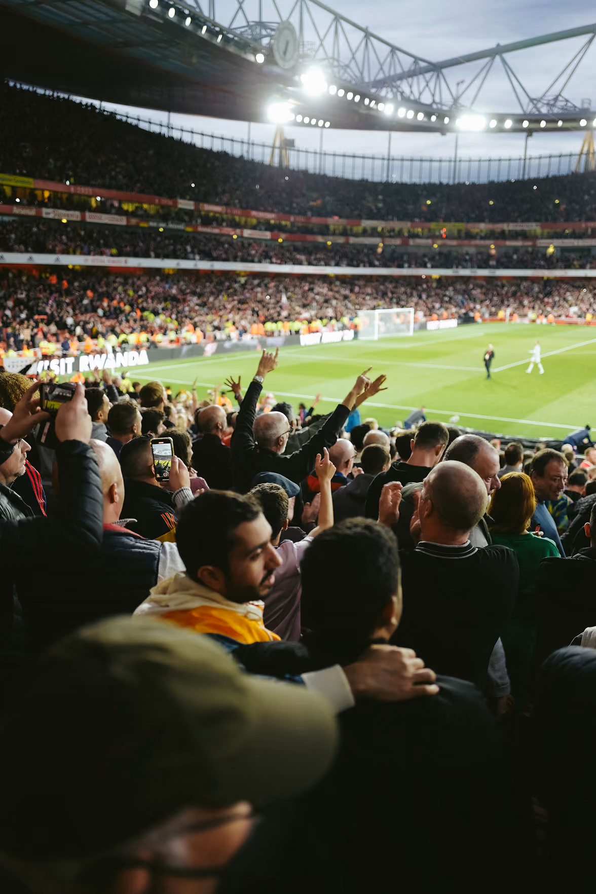 Crowd of enthusiastic soccer fans in a stadium, some raising their hands and taking photos, with a partially visible soccer field and goal in the background under bright stadium lights.