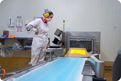 A worker in protective clothing inspects packaging on a conveyor belt in a food processing facility.