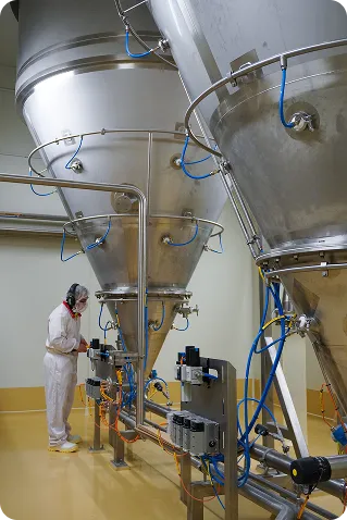 Person in white protective suit and mask operating stainless steel industrial machines with blue tubes in a clean facility.
