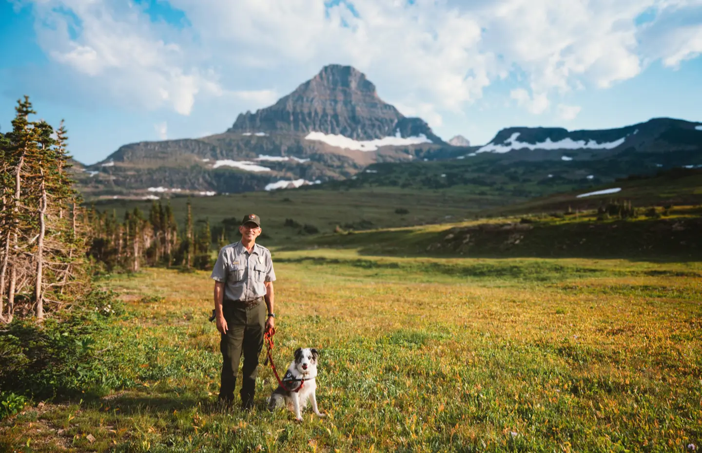 Photo Les Others Magazine - Un homme en uniforme debout avec un chien sur une prairie devant une montagne avec des restes de neige.
