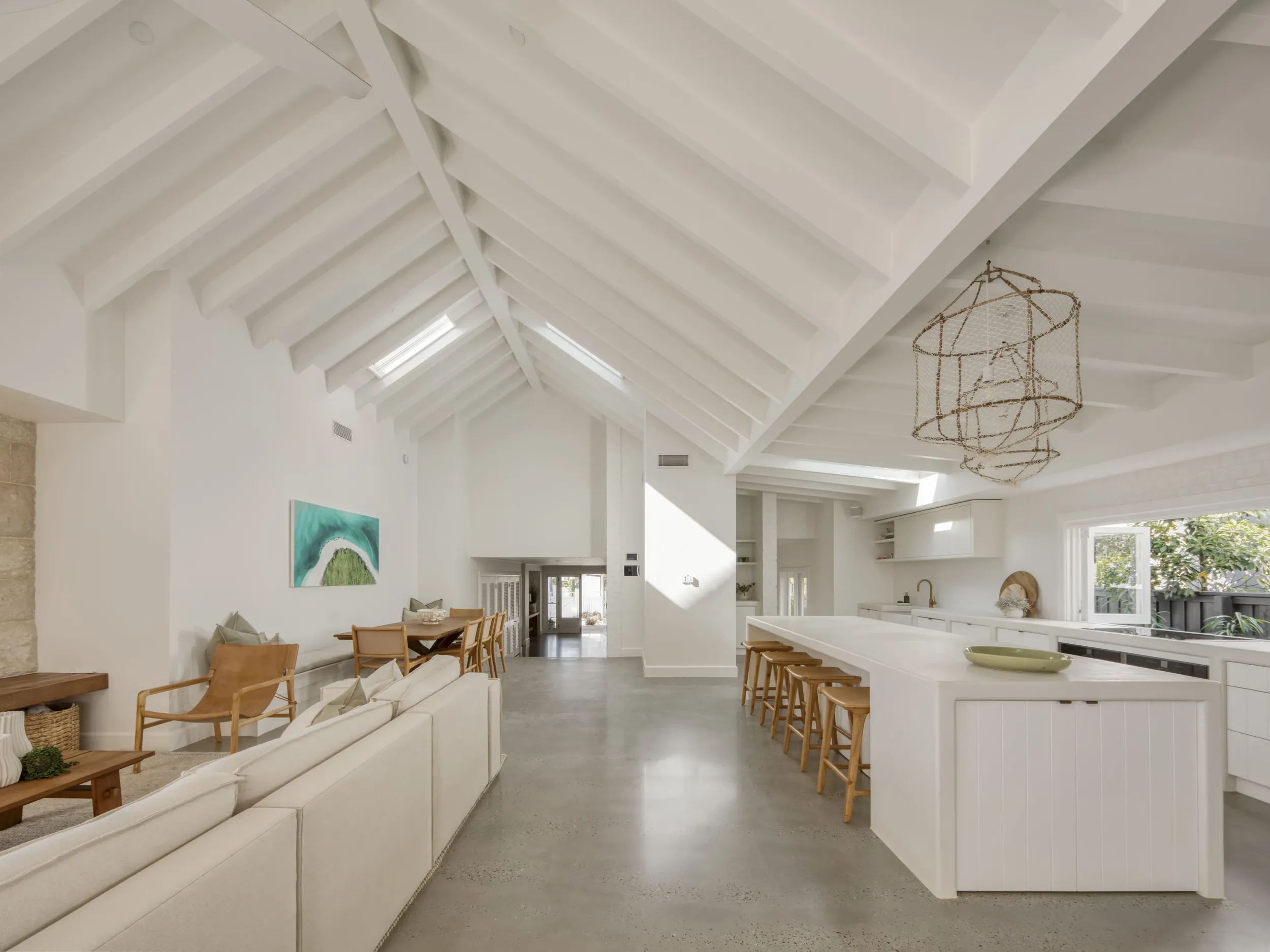 Bright open-concept living space with white vaulted ceiling, skylights, and polished concrete floor; features cream sectional sofa, wooden armchair, dining table with chairs, and large kitchen island with wooden stools under woven pendant lights.